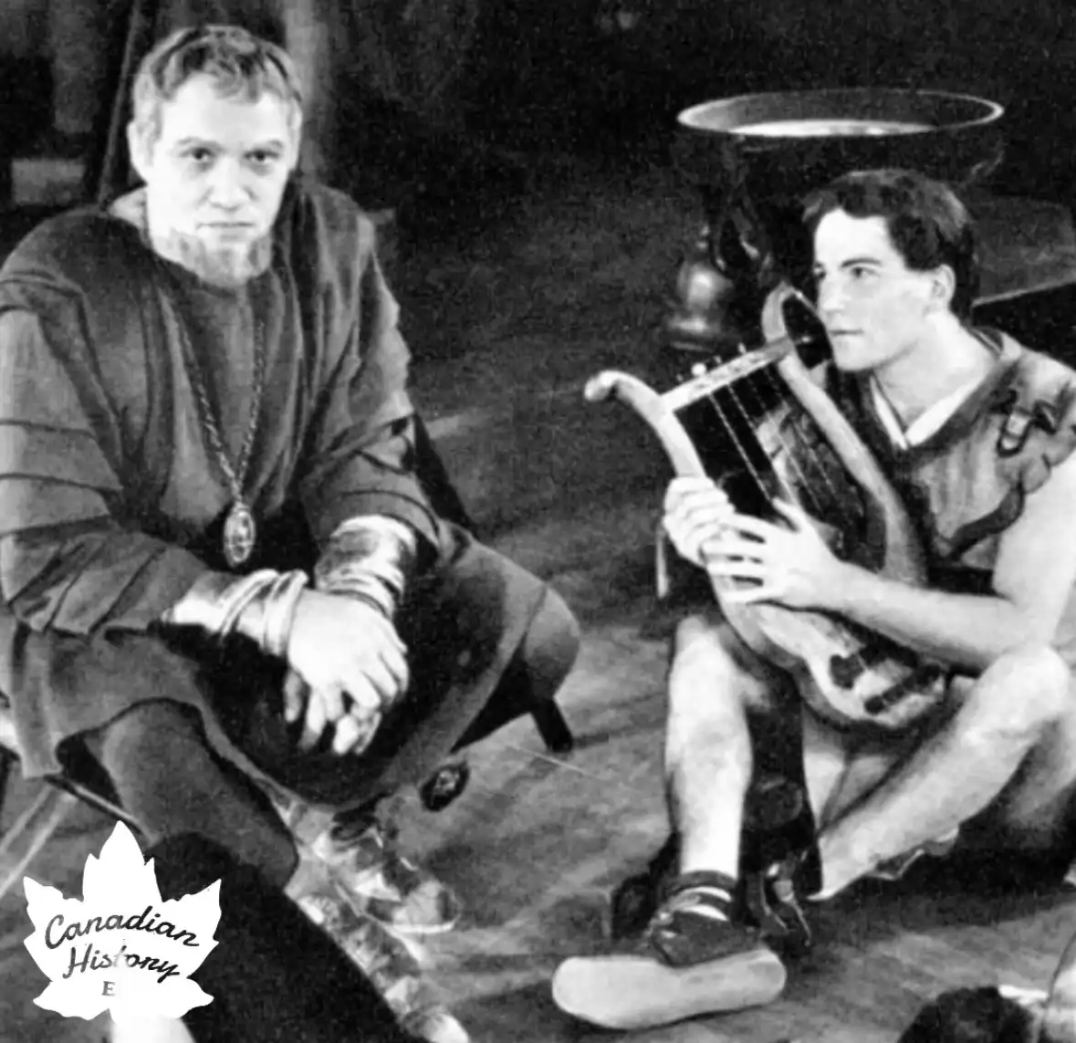 A black-and-white photo shows two men in historical costumes, one seated with a stern expression and the other playing a…