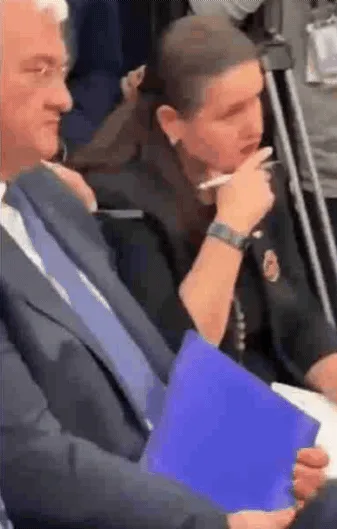 A woman in a dark uniform sits in an audience, holding a blue folder, and uses a white pen to wipe her eye as she appears to…