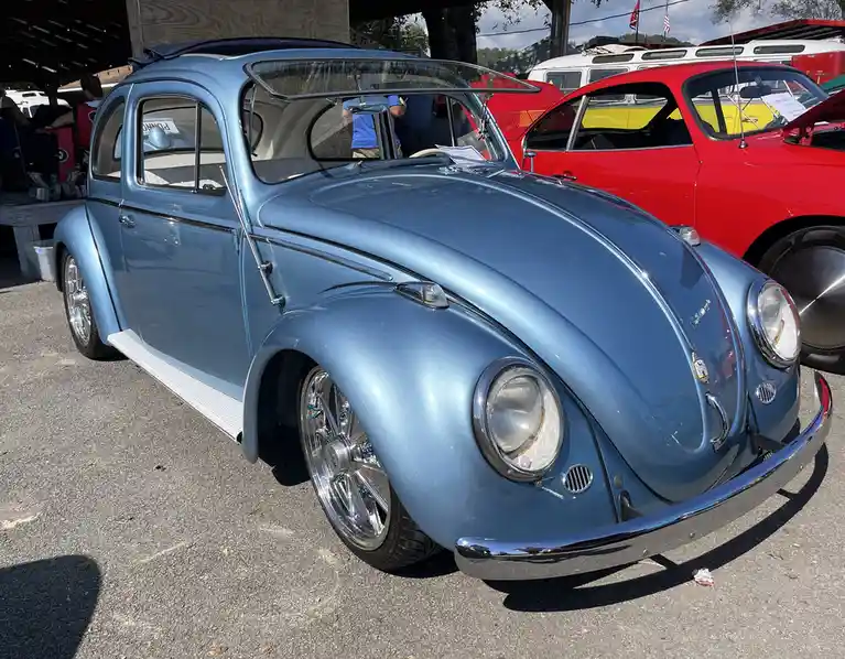 A light blue classic Volkswagen Beetle with chrome accents and custom wheels is parked at an outdoor car show, with a red…