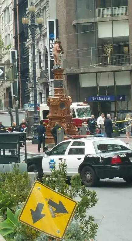 A naked man stands atop a decorative public fountain in a city street, surrounded by police cars, officers, and bystanders…
