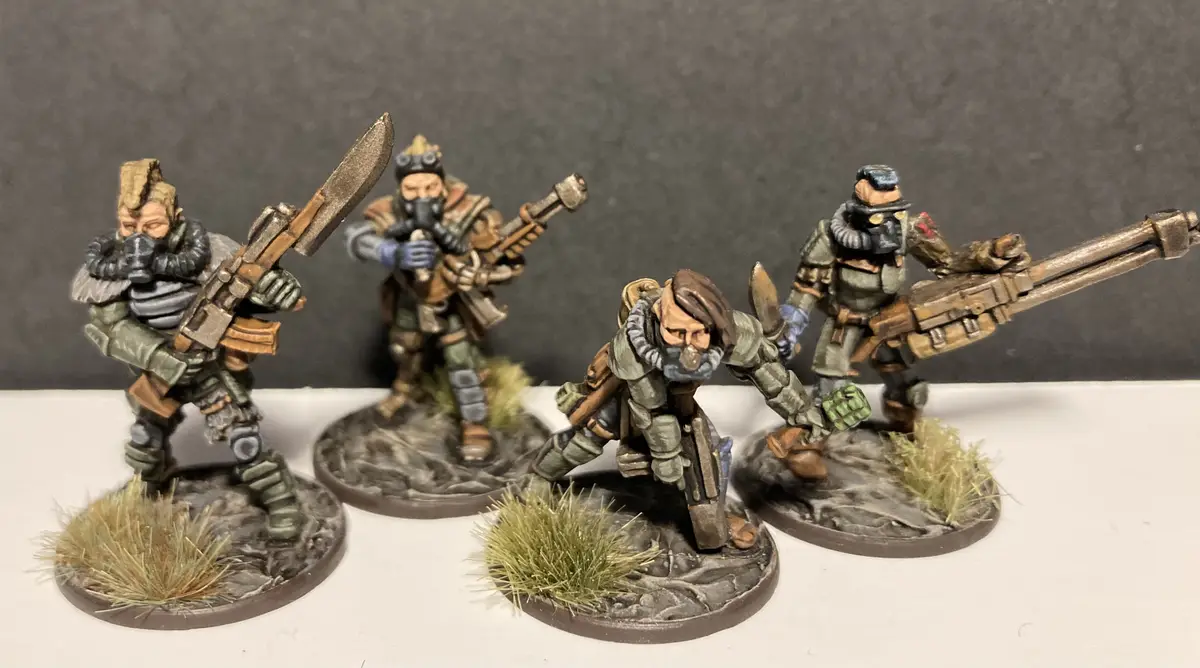 Four painted miniature figures in post-apocalyptic or sci-fi gear, each holding weapons and wearing gas masks or…