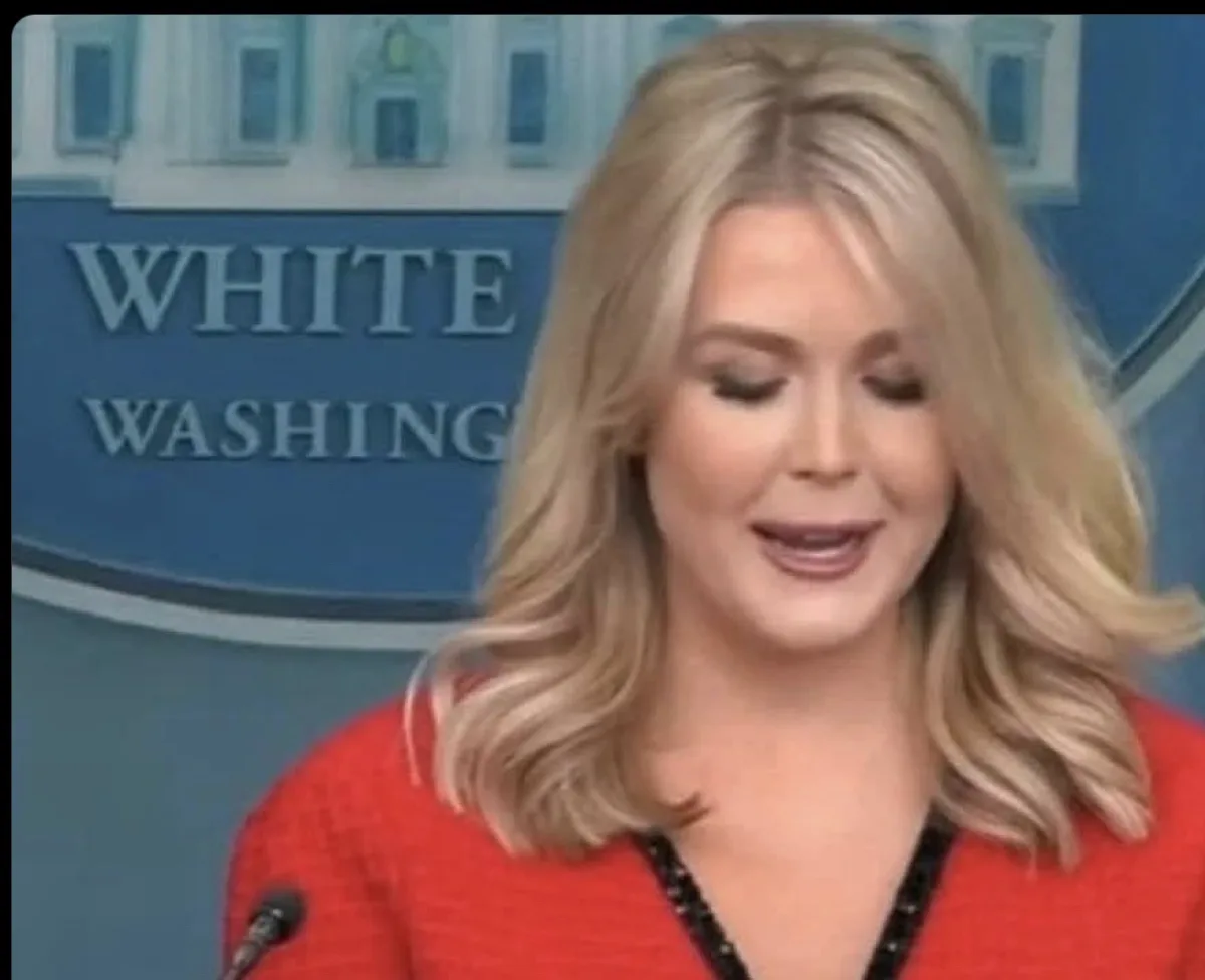 A woman with blonde hair wearing a red top stands in front of a blue backdrop with 'WHITE WASHINGTON' partially visible. She…