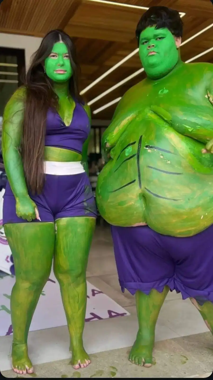 Two people, a woman and a man, are painted entirely green to resemble the Hulk and She-Hulk, wearing purple costumes. The…