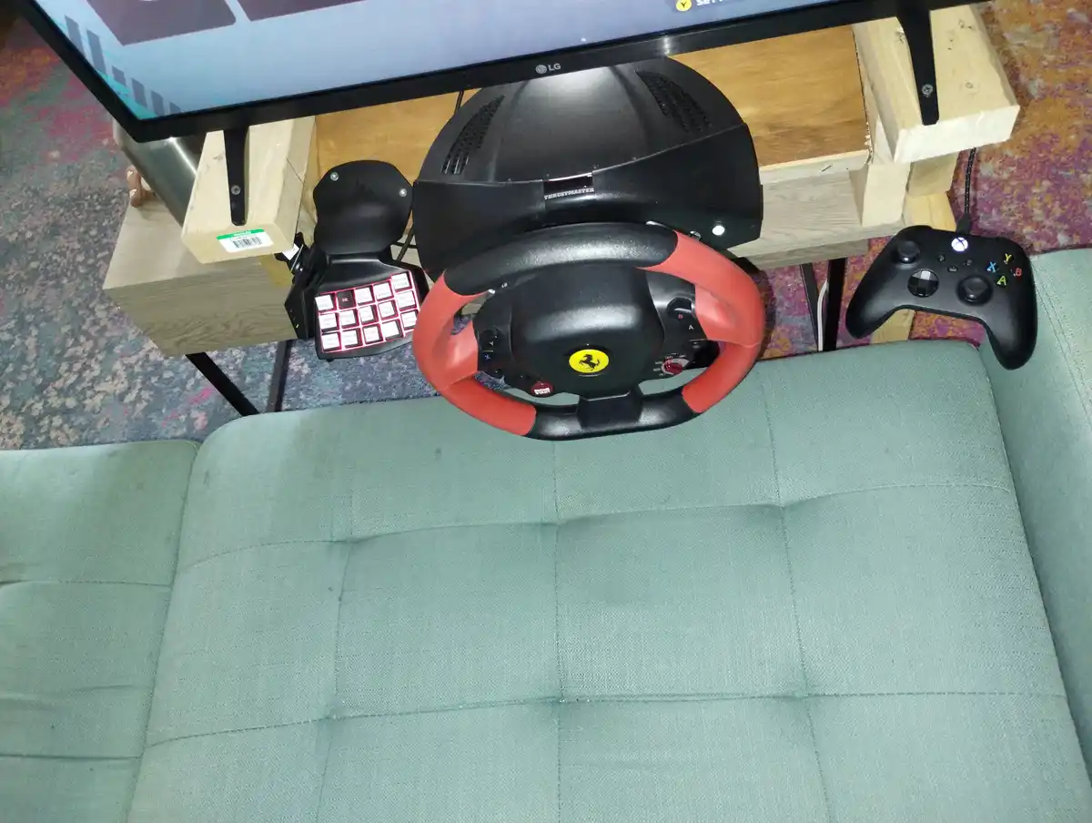 An overhead shot shows a green couch with a Thrustmaster Ferrari racing wheel and pedal set mounted on a wooden stand in…