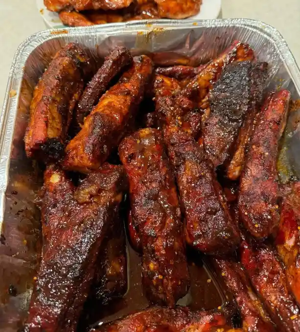 A close-up photo shows a foil tray filled with glossy, saucy, charred ribs, some pieces appearing burnt. Another plate of…