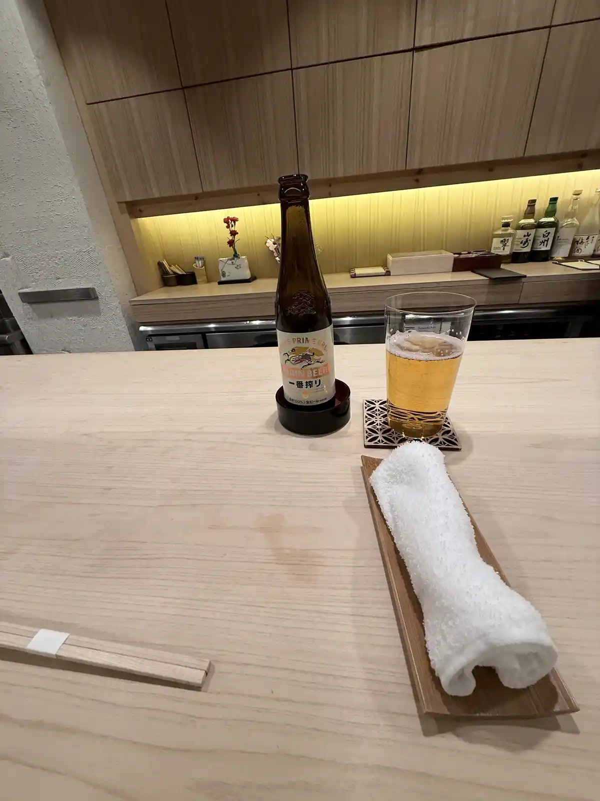 A minimalist Japanese bar scene features a Kirin beer bottle and glass on a light wood counter, alongside a rolled white…