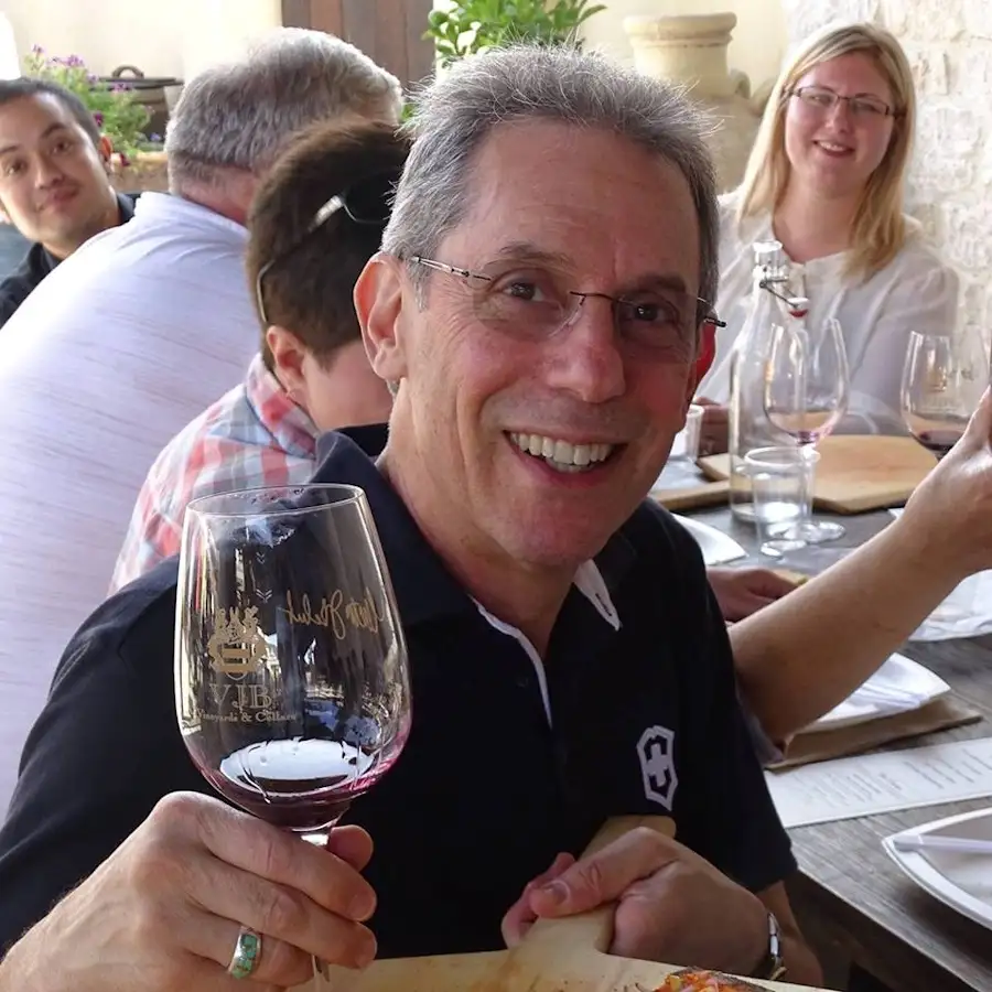 A man with glasses and gray hair smiles broadly while holding up a wine glass with red wine, seated at an outdoor table with…