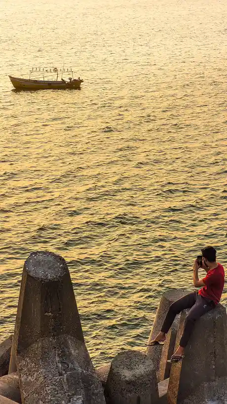 A person in a red shirt sits on a concrete pier, holding up a phone to photograph a small yellow boat on golden-hued water…