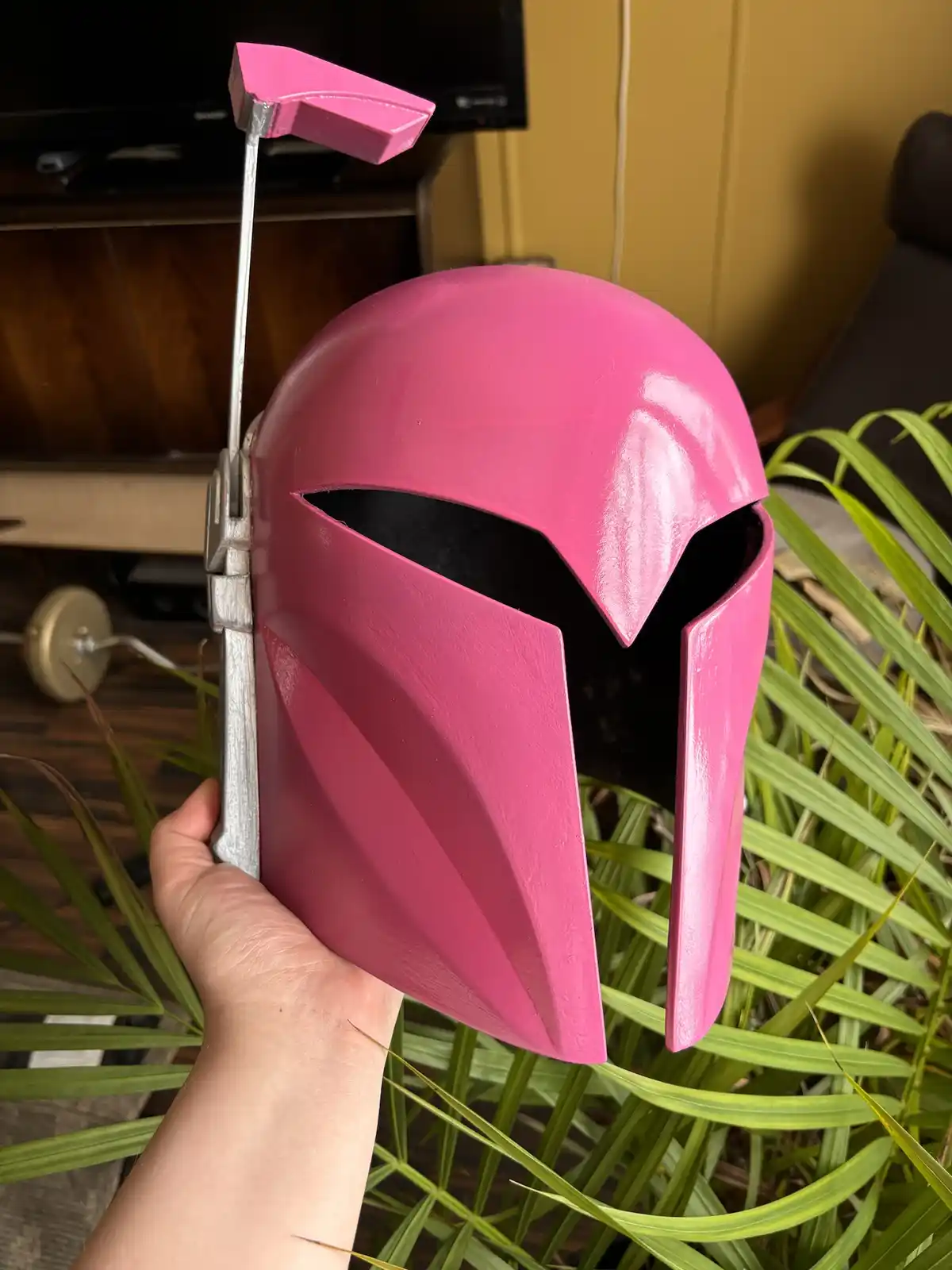 A hand holds a bright pink Mandalorian-style helmet with a silver-colored visor and a pink golf club head attached to the…
