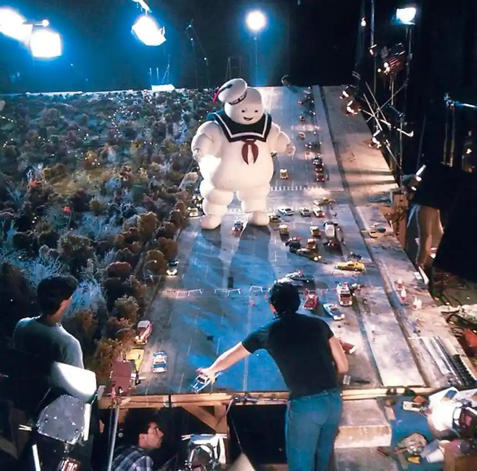 Behind-the-scenes photo from the Ghostbusters movie showing the giant Stay Puft Marshmallow Man prop standing on a miniature…