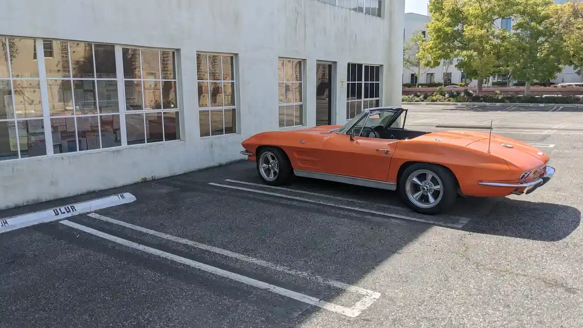 An orange classic convertible sports car is parked in a marked parking space next to a white building with large windows…