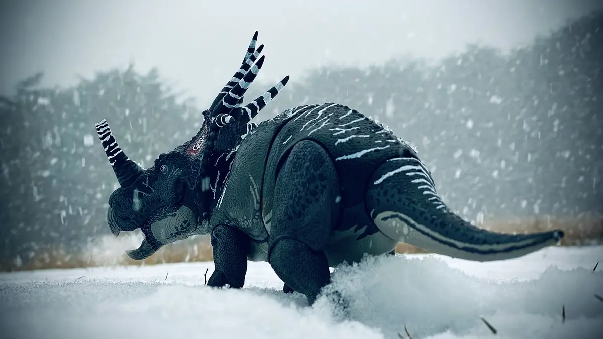 A black and white striped toy triceratops figurine stands in a snowy field with snow falling around it, its horns and body…