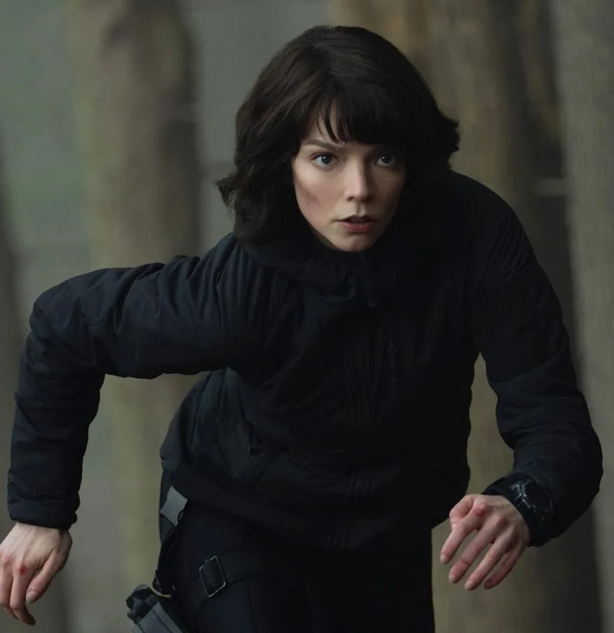 A woman with dark hair and a black jacket is crouched in a tense, alert posture, looking intensely forward. She wears a…