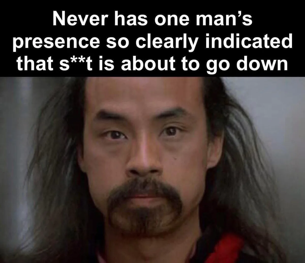 A close-up image of a man with long dark hair and a mustache, looking intensely forward with a serious expression. White…