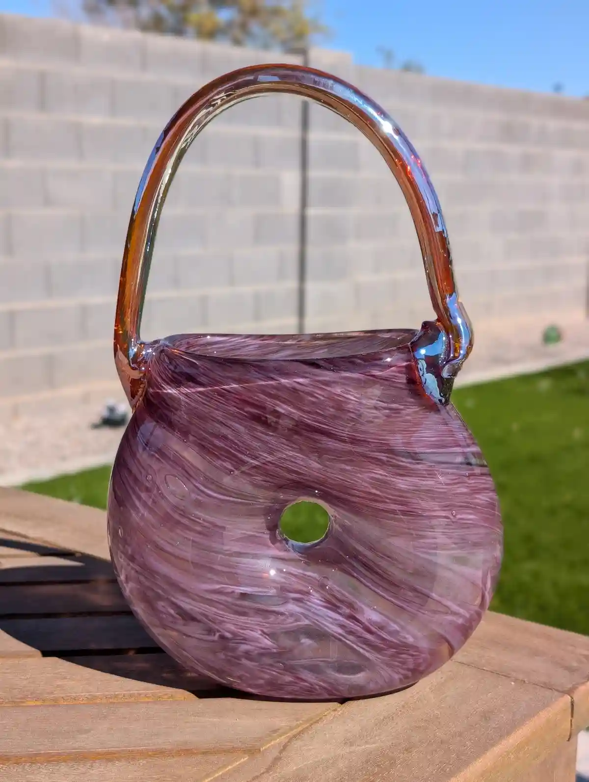 A purple and white swirled glass handbag with a curved handle sits on a wooden table outdoors, featuring a small circular…