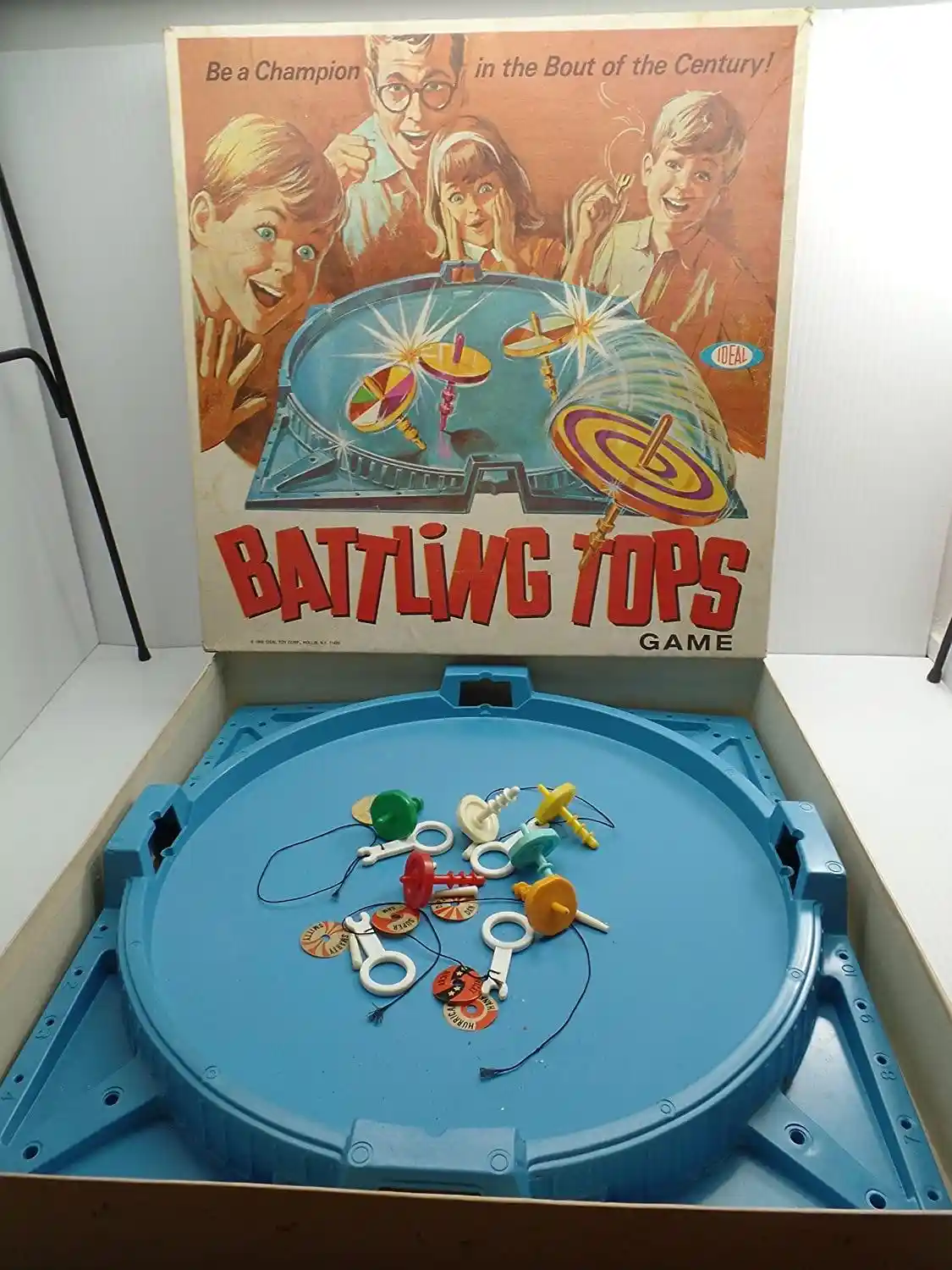 An open vintage 'Battling Tops Game' box shows its blue plastic arena with colorful tops and strings scattered inside. The…
