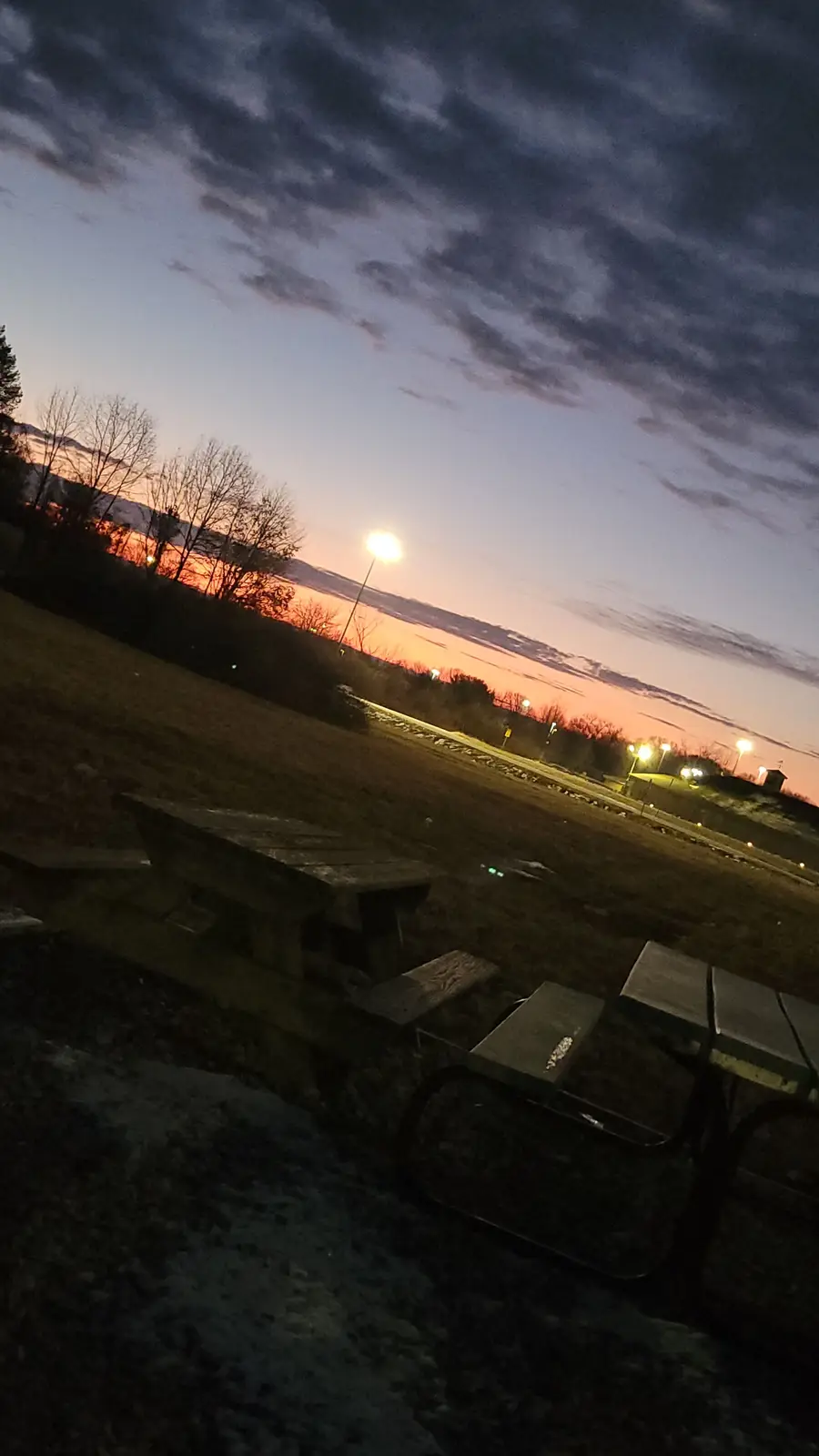 A photo taken at dusk shows a wooden picnic table in the foreground with a dark, snowy ground. In the background, a vibrant…