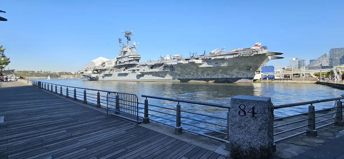 A wide shot shows a large gray aircraft carrier docked in a harbor under a clear blue sky, with a wooden boardwalk and metal…
