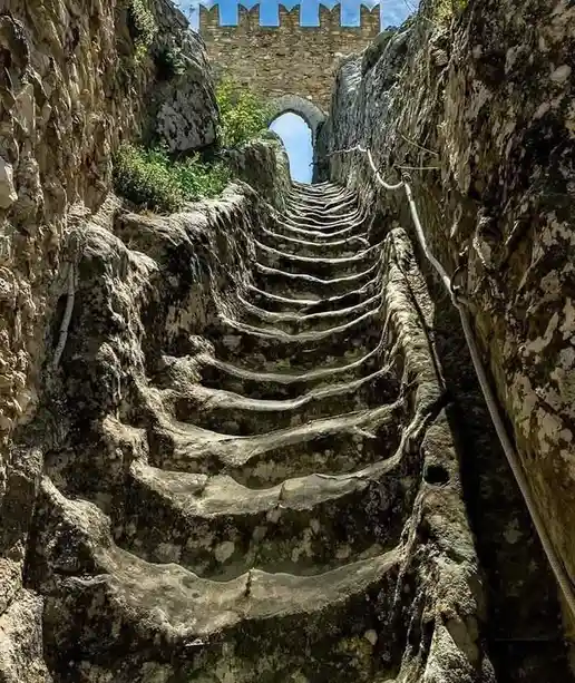 A steep, narrow stone staircase carved into a rocky cliffside, flanked by ancient stone walls and ropes for support, leading…