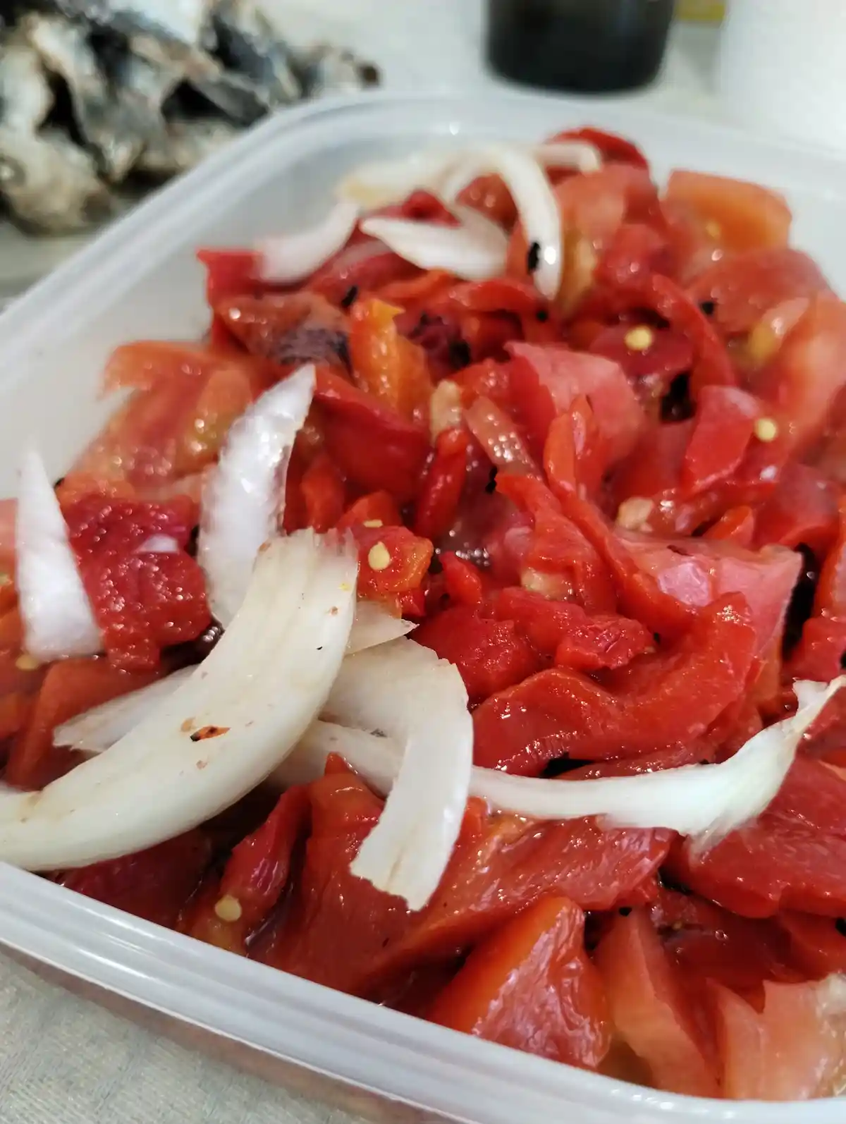 A close-up photo shows a white plastic container filled with chopped red tomatoes and sliced white onions, with some dark…