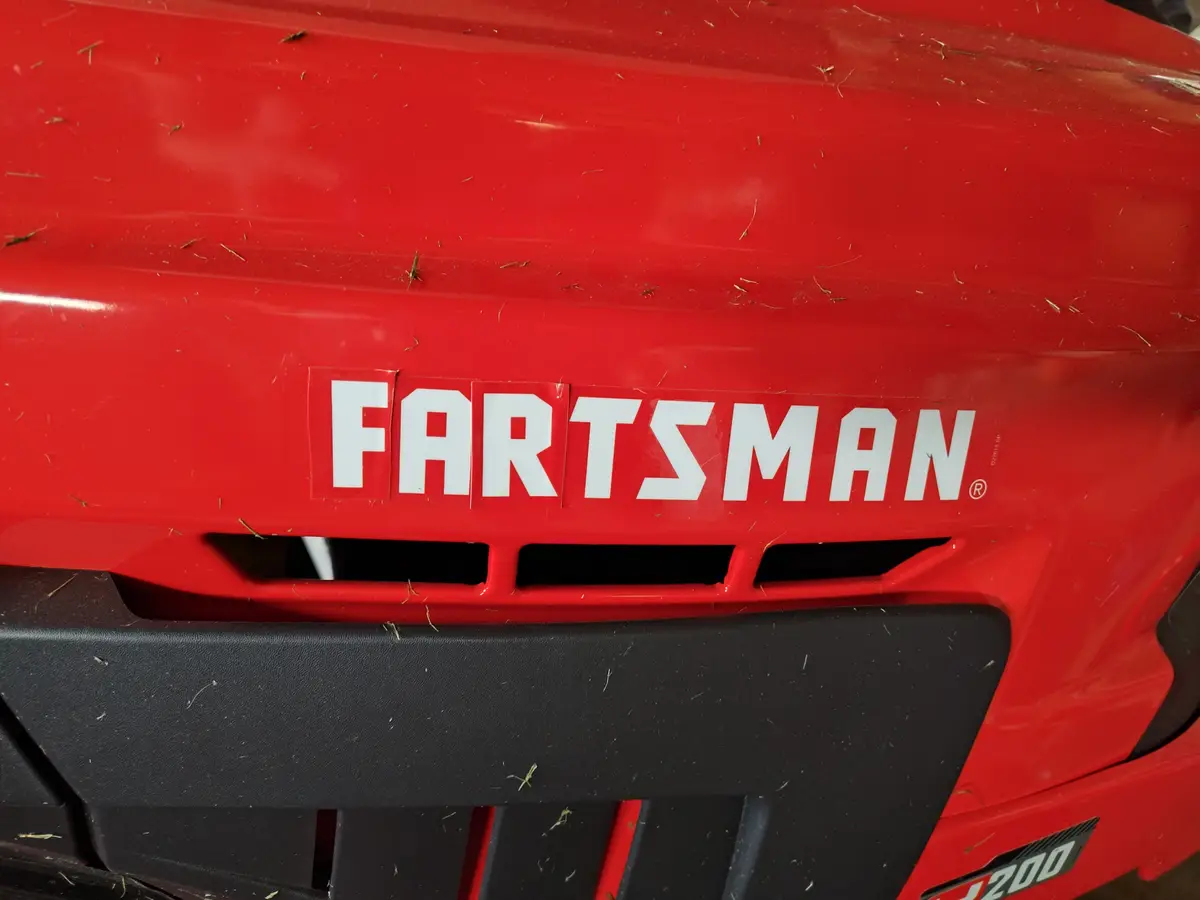 A close-up of a red lawnmower's front grille shows a white sticker with the word 'FARTSMAN' in bold, block letters…