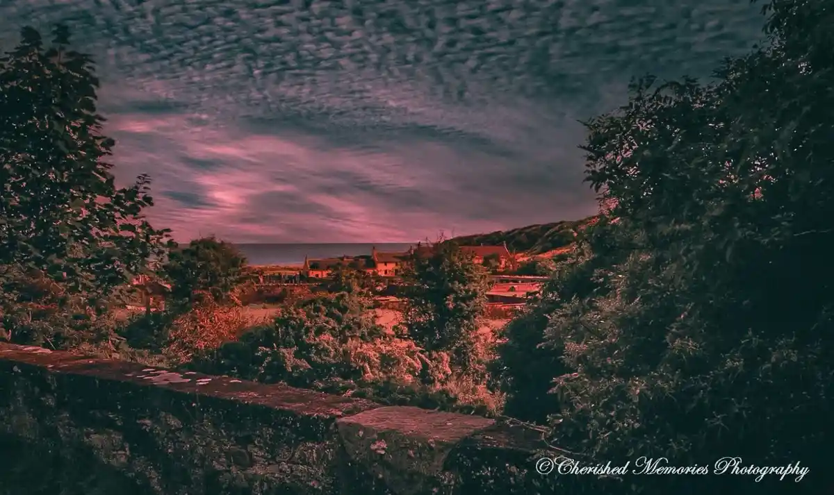 A scenic photograph of a coastal village at dusk, framed by dark trees and a stone wall in the foreground, with a dramatic…
