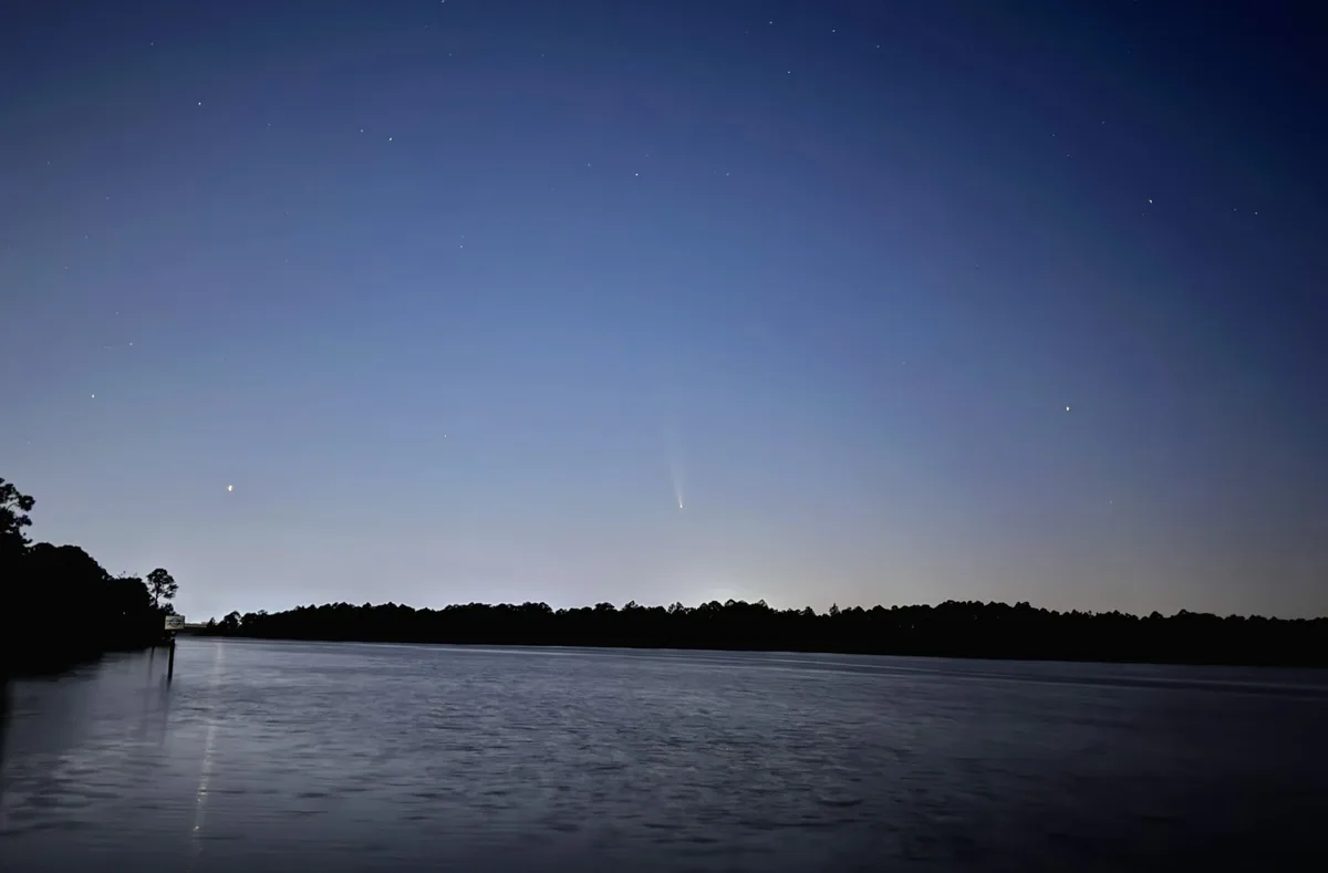 A serene night scene over a calm body of water with a dark treeline in the distance under a starry sky. A bright comet with…