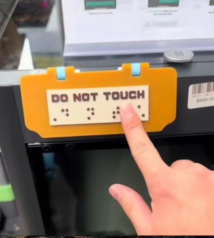 A close-up image shows a person's finger pressing against a 'DO NOT TOUCH' sign that also features braille dots below the…