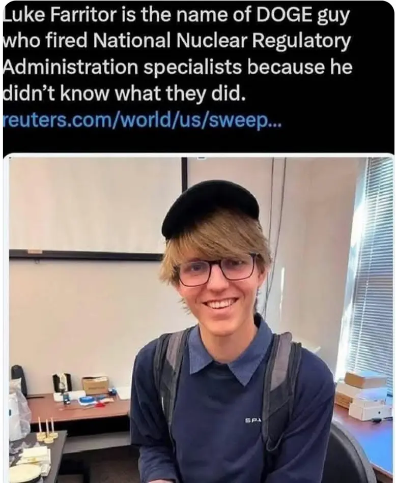 A young man with glasses and a black cap smiles at the camera while wearing a blue shirt and backpack in an office setting…