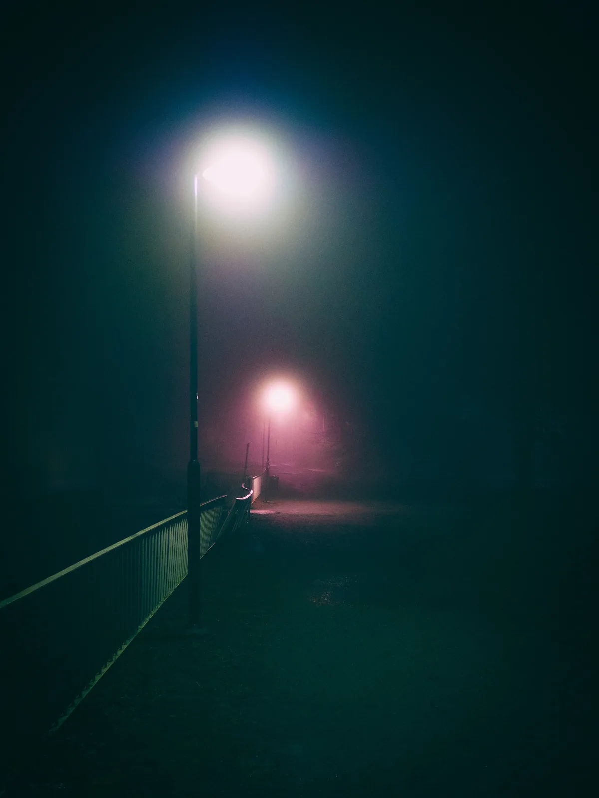 A dark, foggy night scene featuring a pathway with two streetlights. The foreground light casts a bright white glow, while…