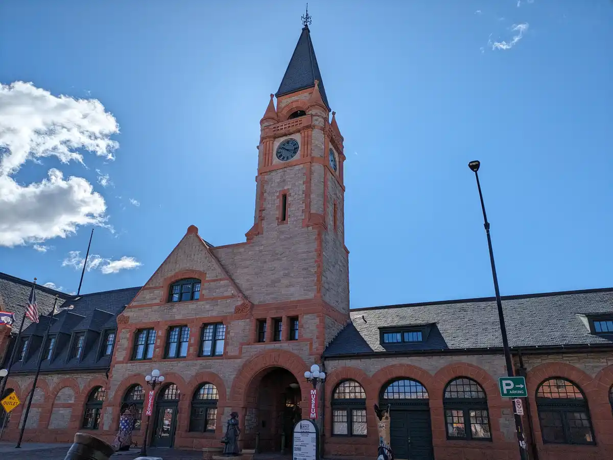 A photo of a grand, historic-looking brick building with a prominent clock tower under a blue sky with white clouds. The…