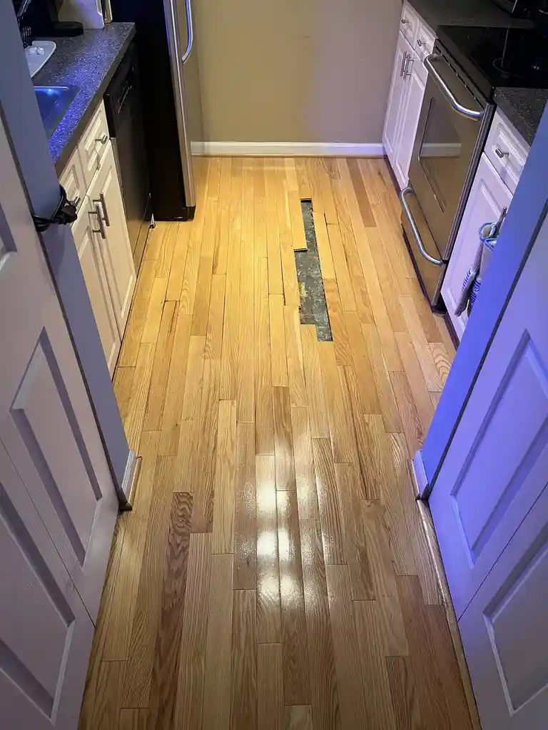 A kitchen with light-colored hardwood flooring has a long, narrow gap in the middle revealing the subfloor, surrounded by…