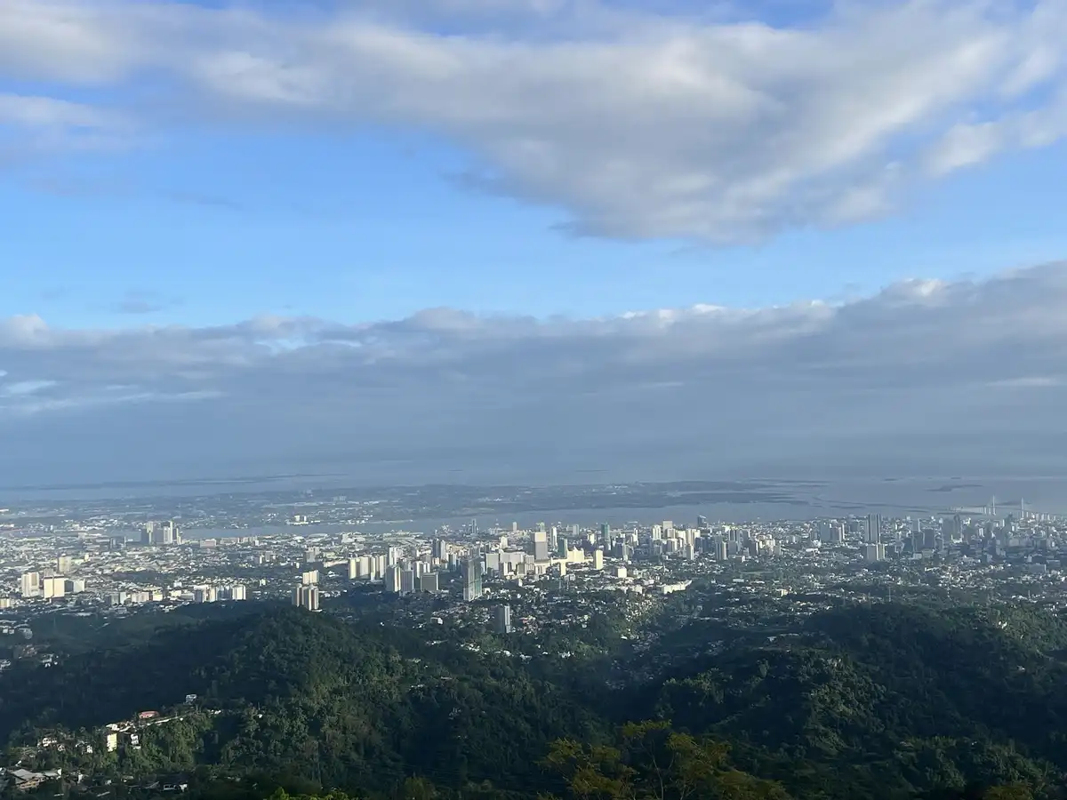A scenic panoramic photo shows a sprawling cityscape with numerous high-rise buildings nestled among green, forested hills…