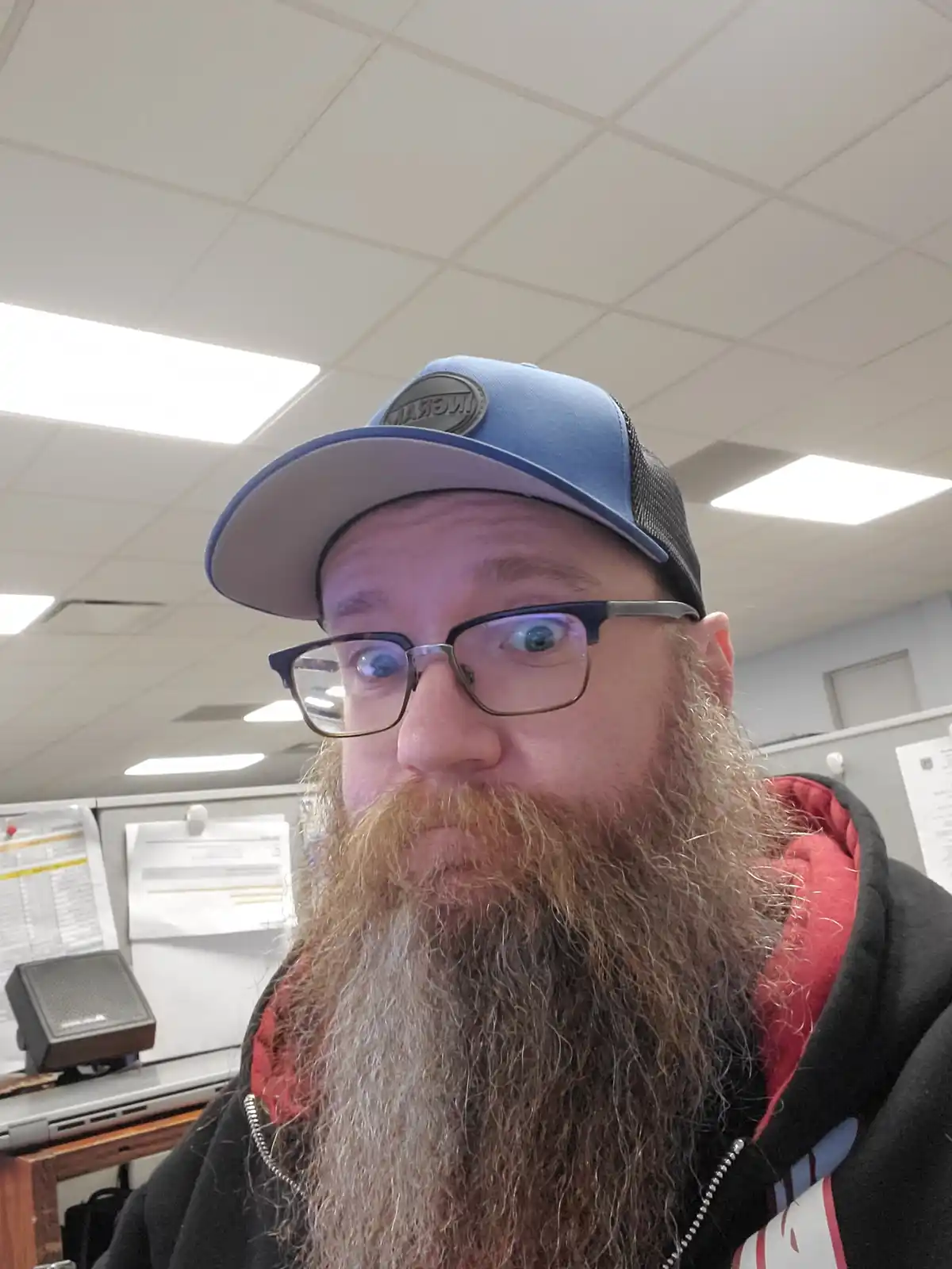 A man with a long, graying beard and glasses wears a blue and black trucker cap while looking directly at the camera with…