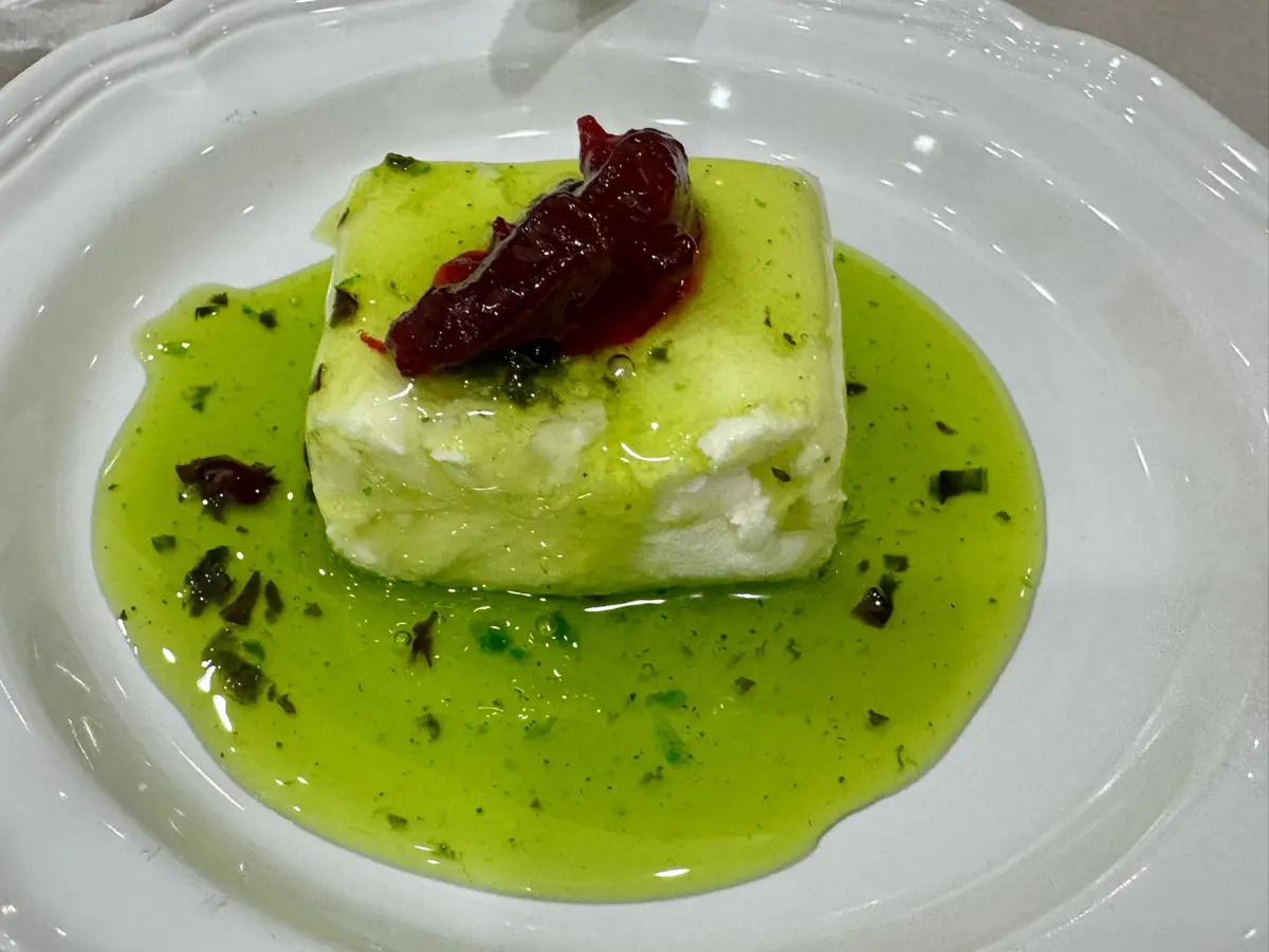 a close-up of a white plate holding a square block of white cheese or tofu, partially submerged in a bright green sauce with…