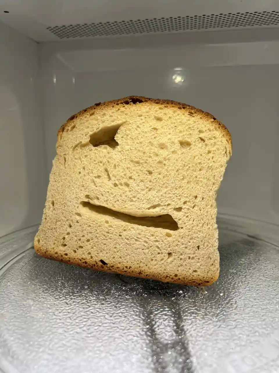 A single slice of bread sits upright inside a microwave, its surface showing two holes that resemble a smiling face. The…