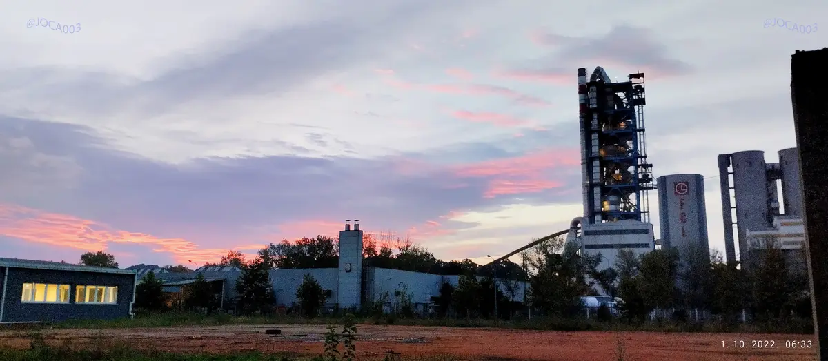 A photograph of an industrial cement plant at dawn, with pink and purple clouds in the sky, and a small building with lit…