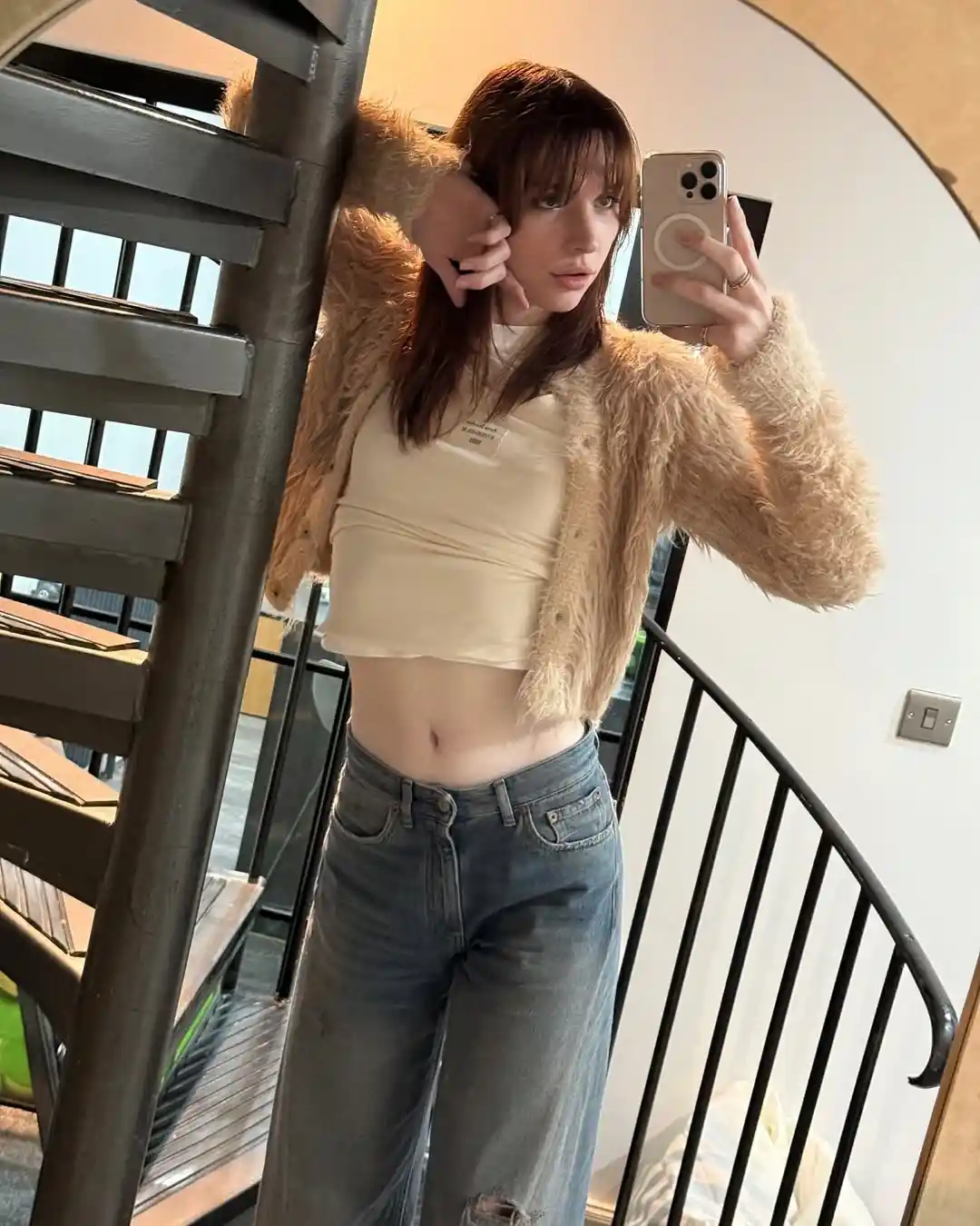 A young woman with brown hair takes a mirror selfie while wearing a beige crop top, a fluffy tan jacket, and ripped jeans…