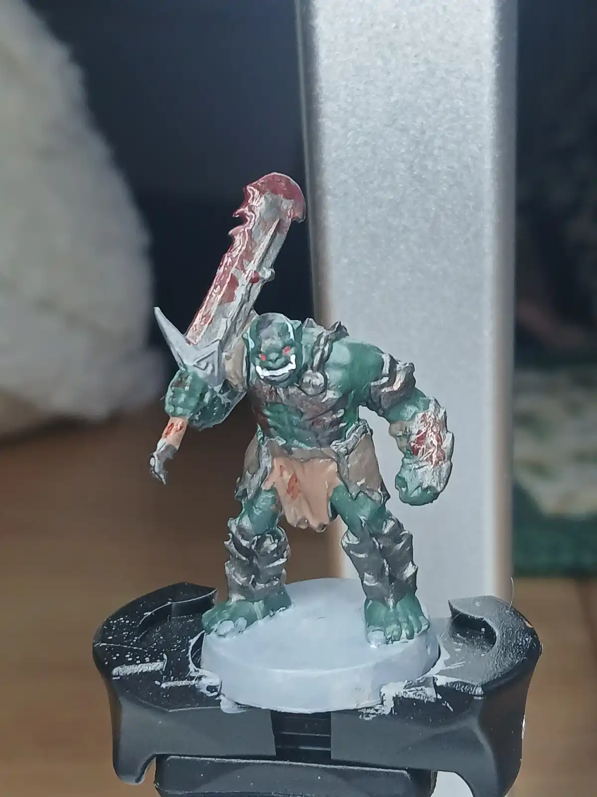 A painted green orc-like miniature figure with red eyes, wielding a large blood-stained sword, stands on a white circular…