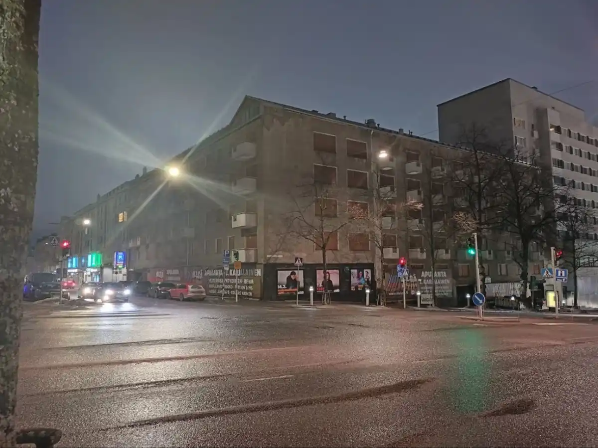 A nighttime street scene showing a wet asphalt road with cars and traffic lights, flanked by multi-story buildings with…