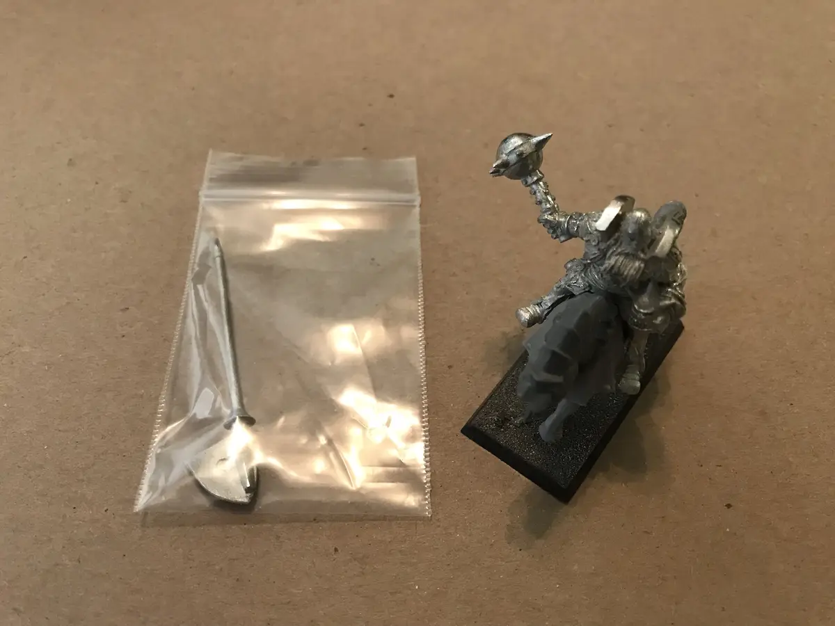 A silver-painted miniature knight figurine on a black base sits next to a clear plastic bag containing a single metal nail…