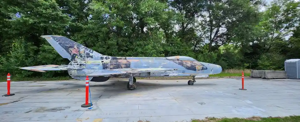 A weathered, rusted military jet with peeling paint and a faded red star on its tail is parked on a concrete pad surrounded…