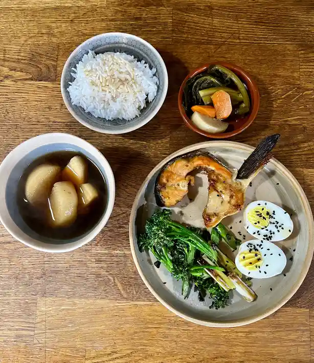 An overhead shot displays a traditional Japanese-style meal on a wooden table, featuring a plate with grilled fish, steamed…