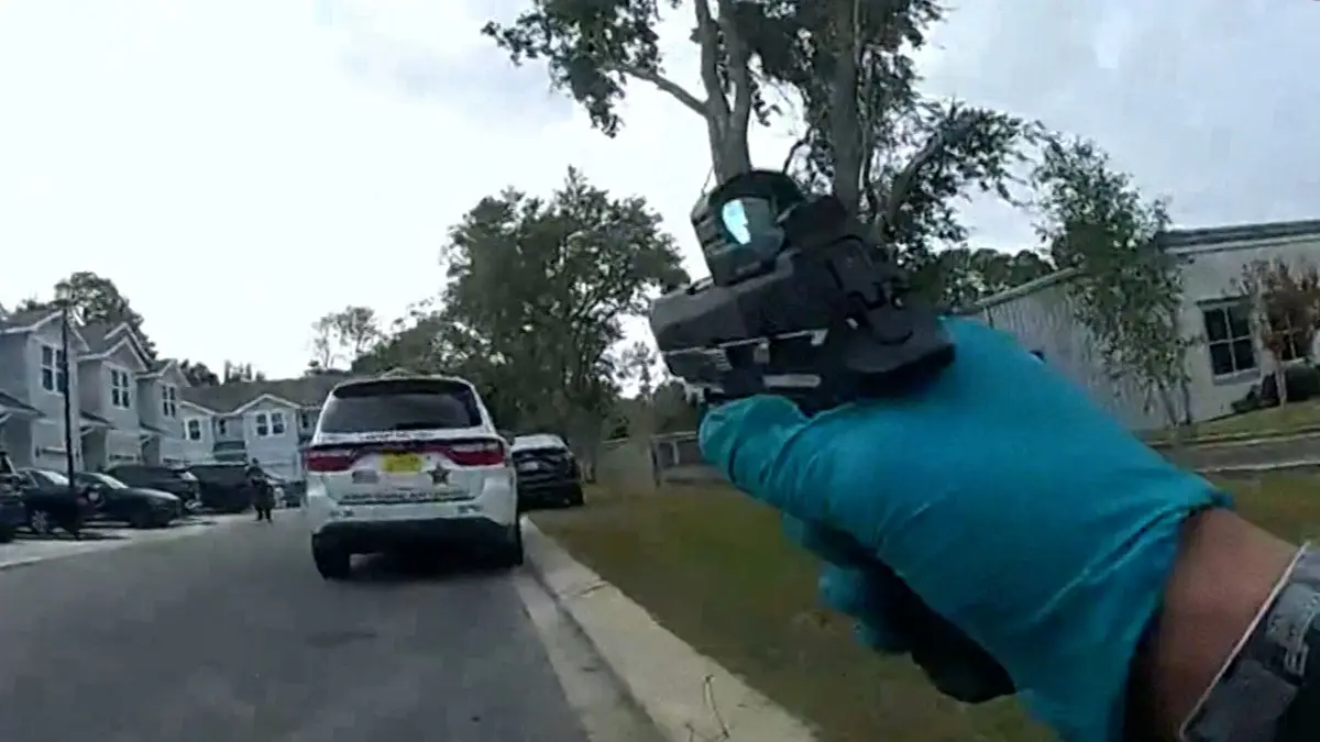 A first-person view shows a gloved hand holding a handgun with a red dot sight, pointed forward down a residential street…