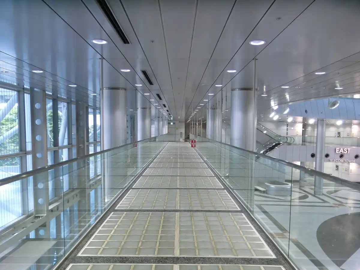 A long, empty, modern glass-walled walkway in a building with 'EAST TELECOM TOWER' signage visible on the right. The floor…