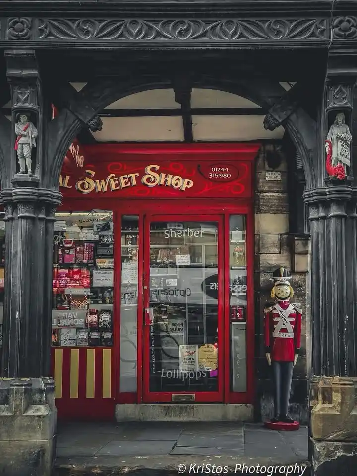 A vintage-style sweet shop with a bright red facade and ornate black stone archway. The glass door and windows display candy…