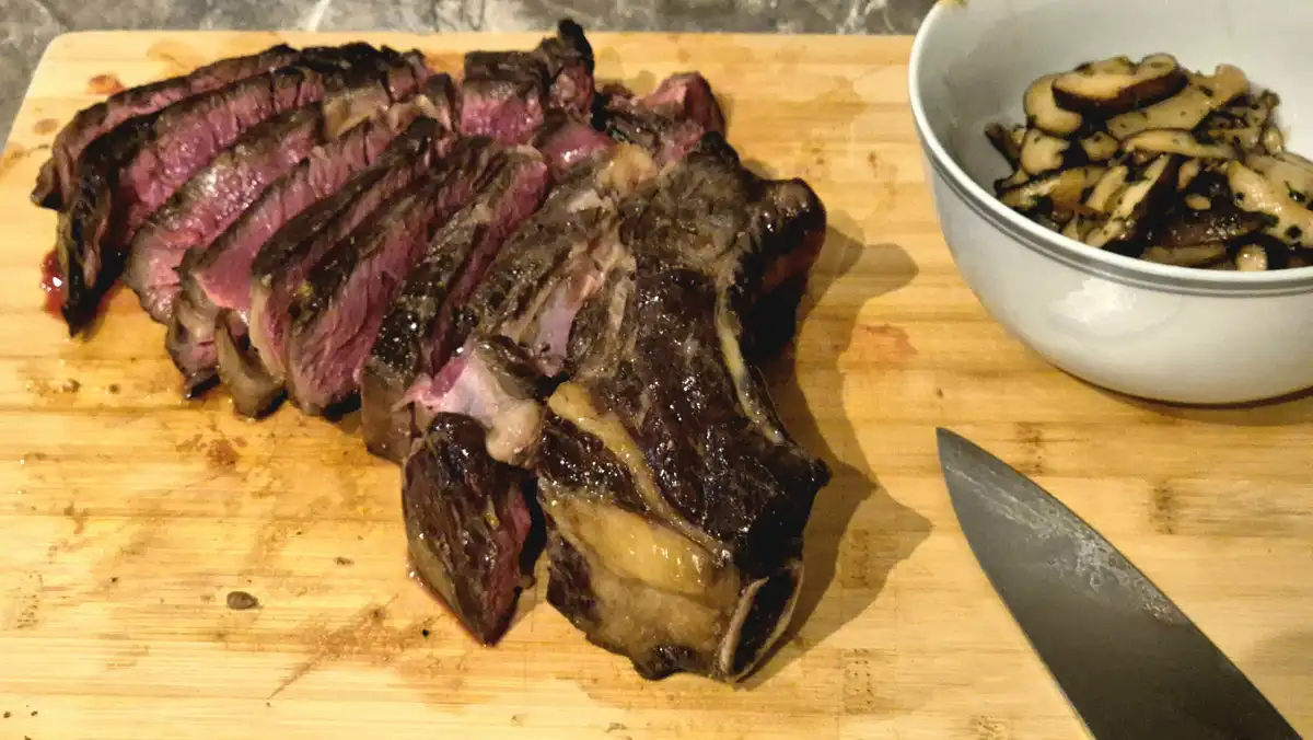 A wooden cutting board holds a sliced, juicy medium-rare steak with a charred exterior, next to a white bowl of sautéed…