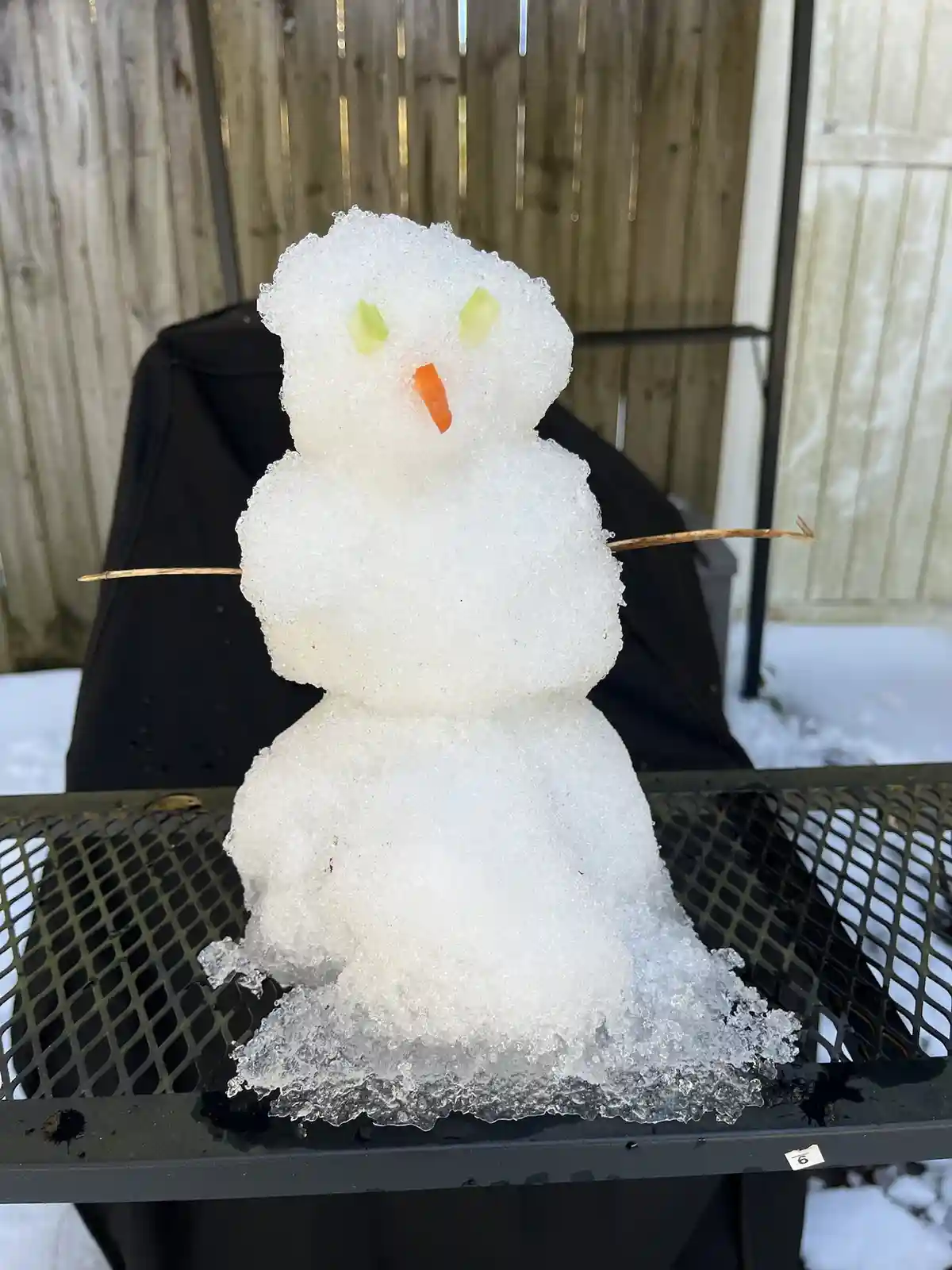A small, three-tiered snowman made of ice sits on a black metal mesh table outdoors, with a wooden fence in the background…