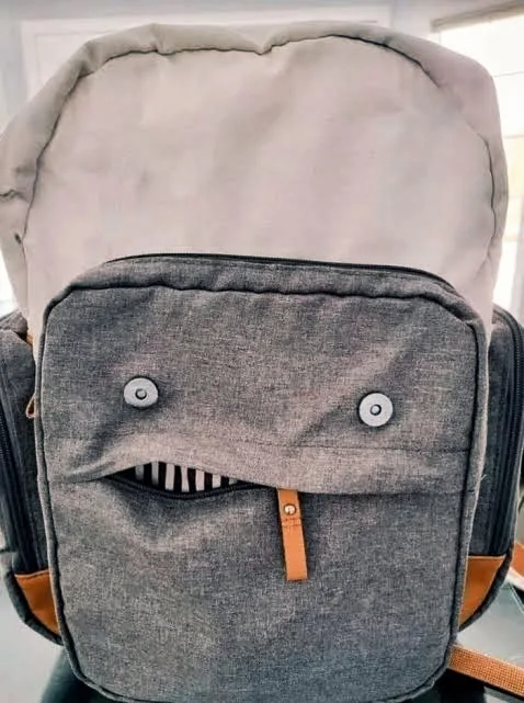 A gray backpack with a zipper pocket that looks like a smiling face, featuring two silver snap buttons as eyes and a zipper…