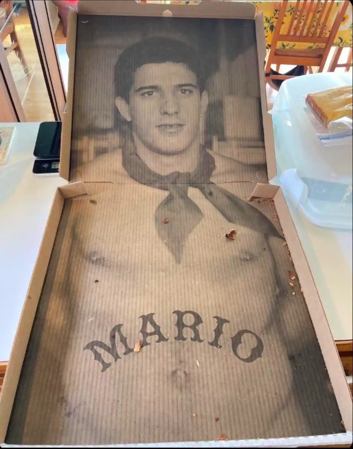 An open cardboard pizza box has a faded black-and-white photo of a young man wearing a neckerchief printed on its inner lid…